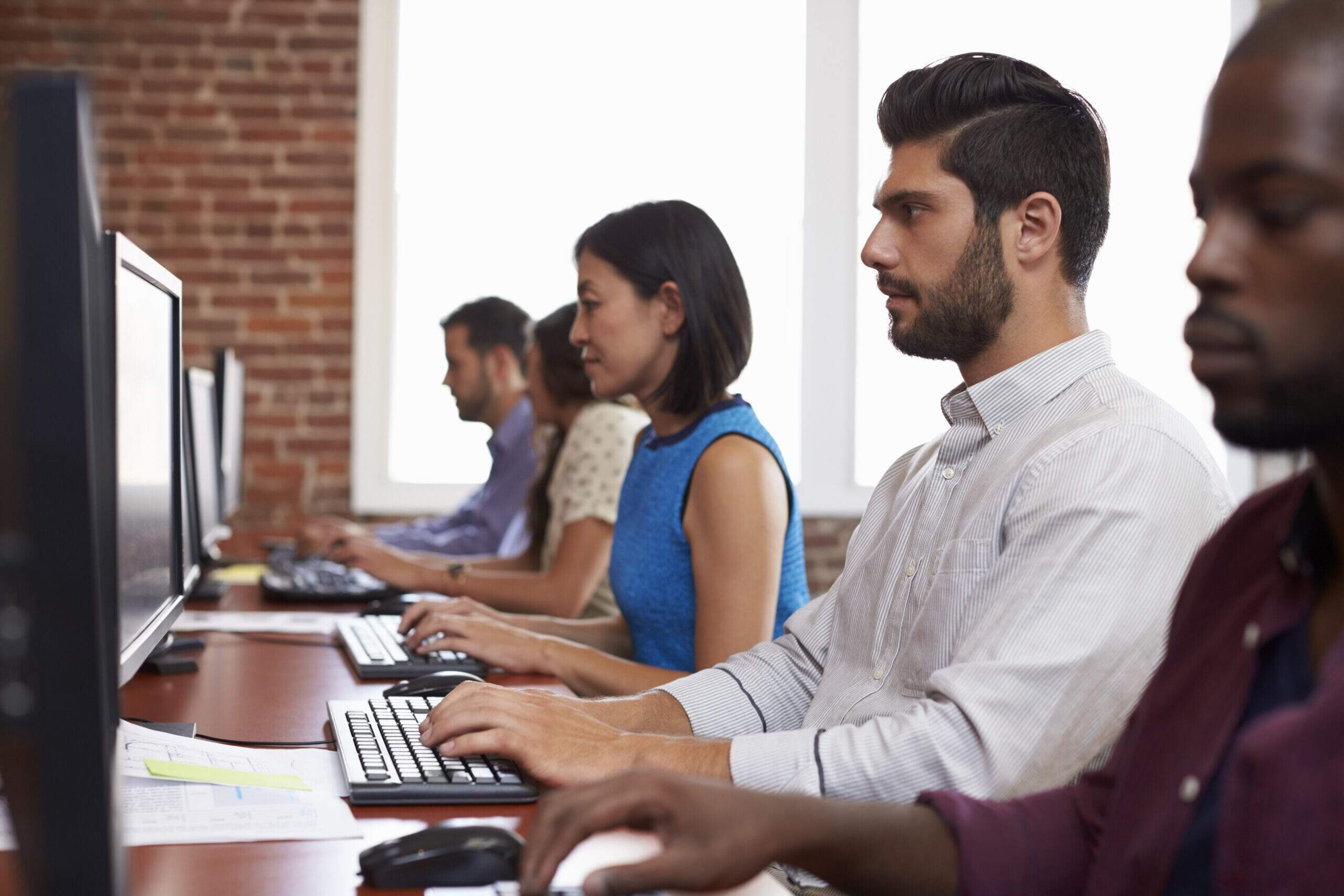 Grupo de pessoas em frente a computadores em ambiente corporativo, com expressões sérias e concentradas, ilustrando a rotina de trabalho e possíveis impactos na saúde mental.